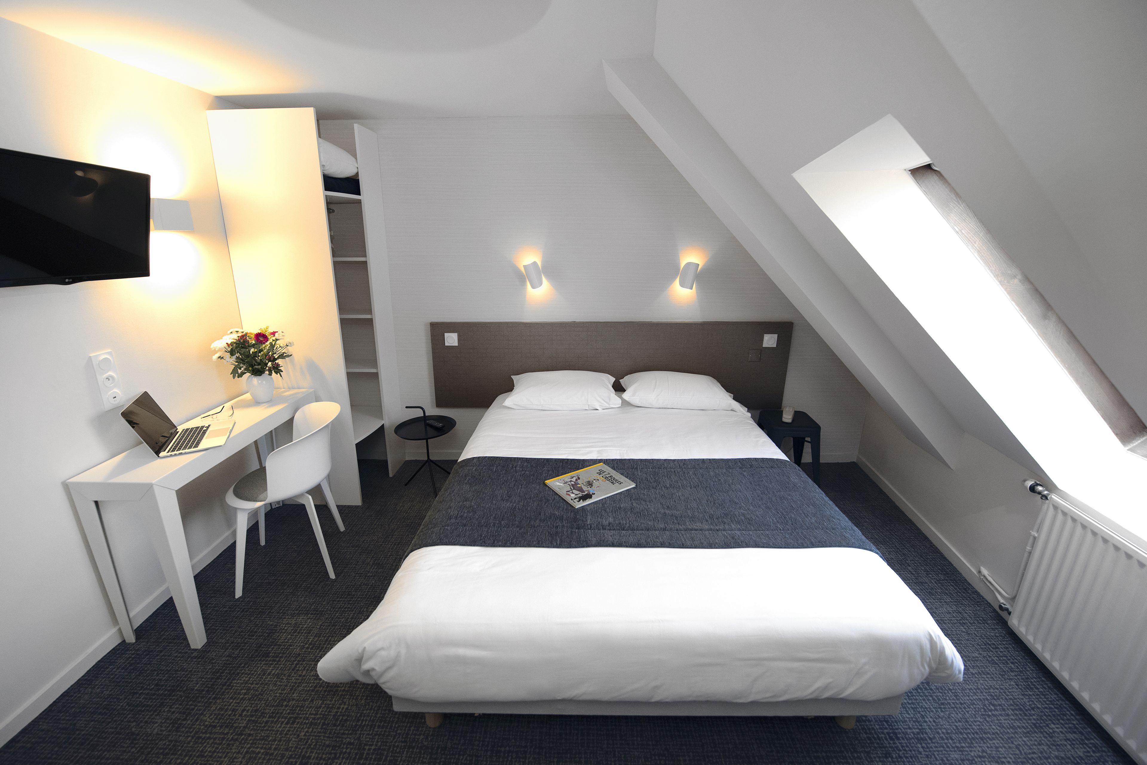 A modern hotel room with a bed, desk, and chair, a flat-screen TV, and a skylight.