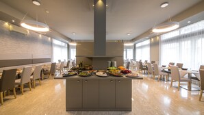 A spacious dining area with multiple tables and chairs, a central buffet island with various food items, and large windows providing natural light. - Hotel Torino (Jesolo)