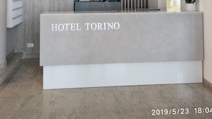 A modern hotel reception desk with a chandelier, a wooden panel wall, and a reception counter. - Hotel Torino (Jesolo)