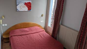 Une chambre à coucher avec une tête de lit en bois, une couvre-lit rouge à motifs, un tableau représentant une rose blanche et rouge accroché au mur, une fenêtre avec des rideaux et un radiateur.
