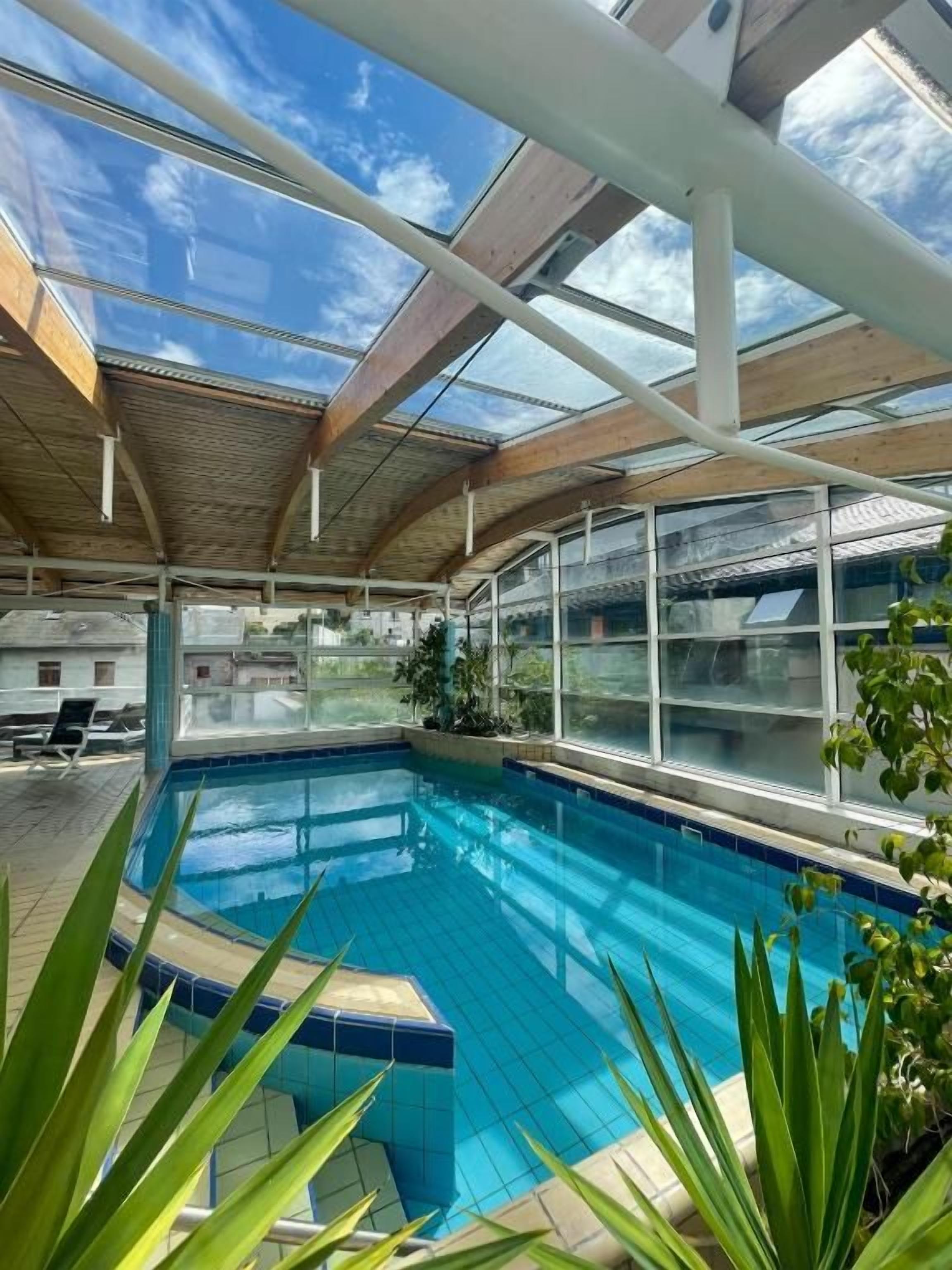 An indoor swimming pool with clear glass ceilings and wooden beams, surrounded by potted plants.
