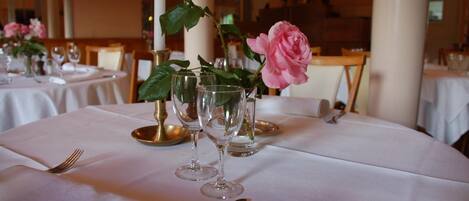 A dining table set with white tablecloths, clear glassware, and a pink rose centerpiece.
