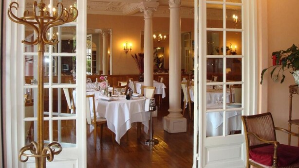 An elegant dining area with white tables and chairs, a chandelier, and a mirror reflecting the room. - Hôtel Régina (La Bourboule)