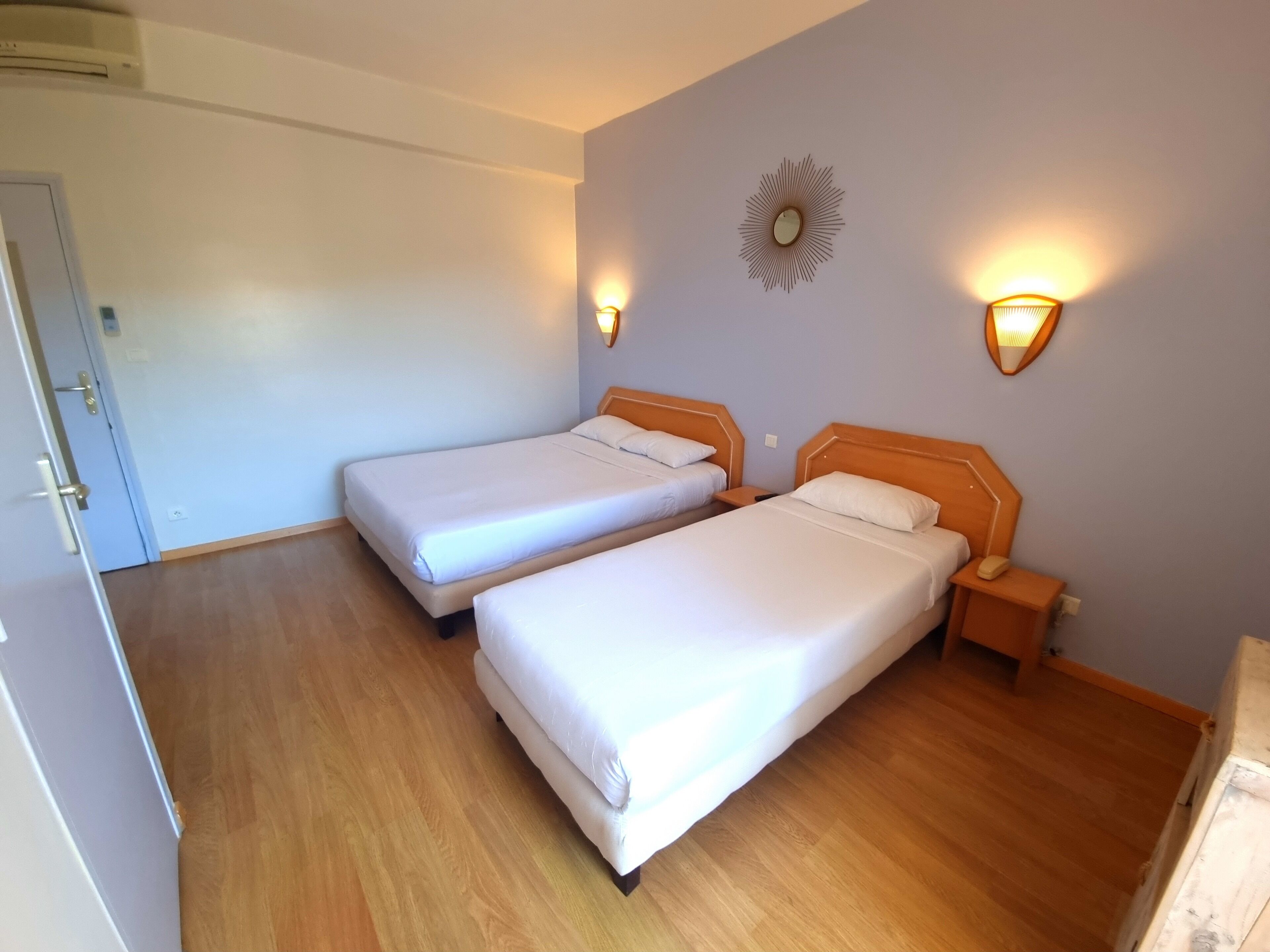 Habitación con dos camas, piso de madera, una puerta e iluminación en la pared.