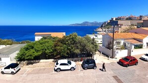 Un pueblo costero con vistas despejadas al mar y montañas en la distancia. Se ven varios coches aparcados y un peatón caminando por la acera.