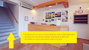 Zona de recepción de un hotel con una escalera, un tablón de anuncios con varias fotografías y un mostrador de madera.