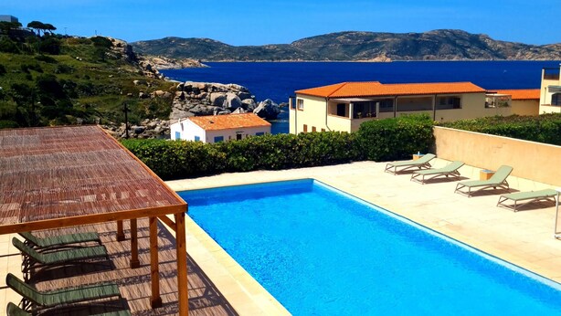 Una piscina con sillas de descanso, una mesa de madera y vistas al mar y las colinas.