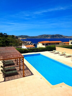 A swimming pool with lounge chairs, a wooden table, and a view of the sea and hills. - Hôtel Sole Mare (Calvi)