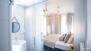 A modern hotel room with a glass partition separating the bathroom and bedroom. The bathroom features a white sink and a mirror. The bedroom has a bed with a beige blanket and pillows, a wooden nightstand, and a chandelier. - Hôtel Le Rapp (Colmar)