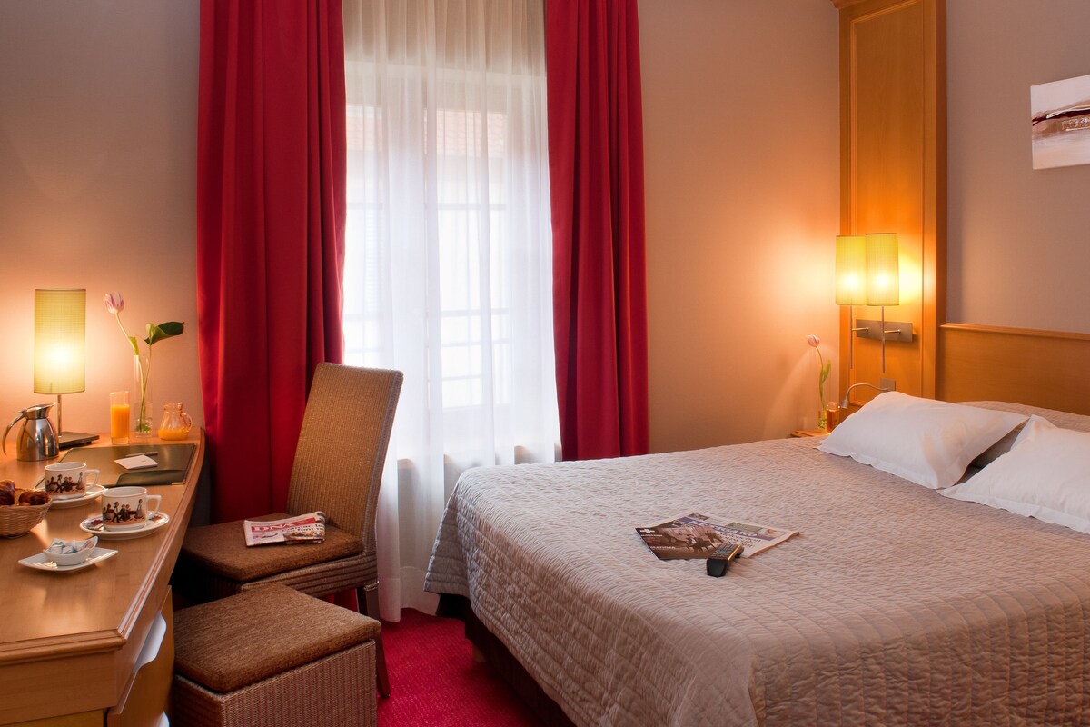 A hotel room with a bed, a desk with breakfast items, a chair, and a window with red curtains.