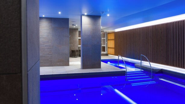 A modern indoor pool area with a blue-lit pool, a wooden wall, and a seating area in the background. - Hôtel Le Rapp (Colmar)