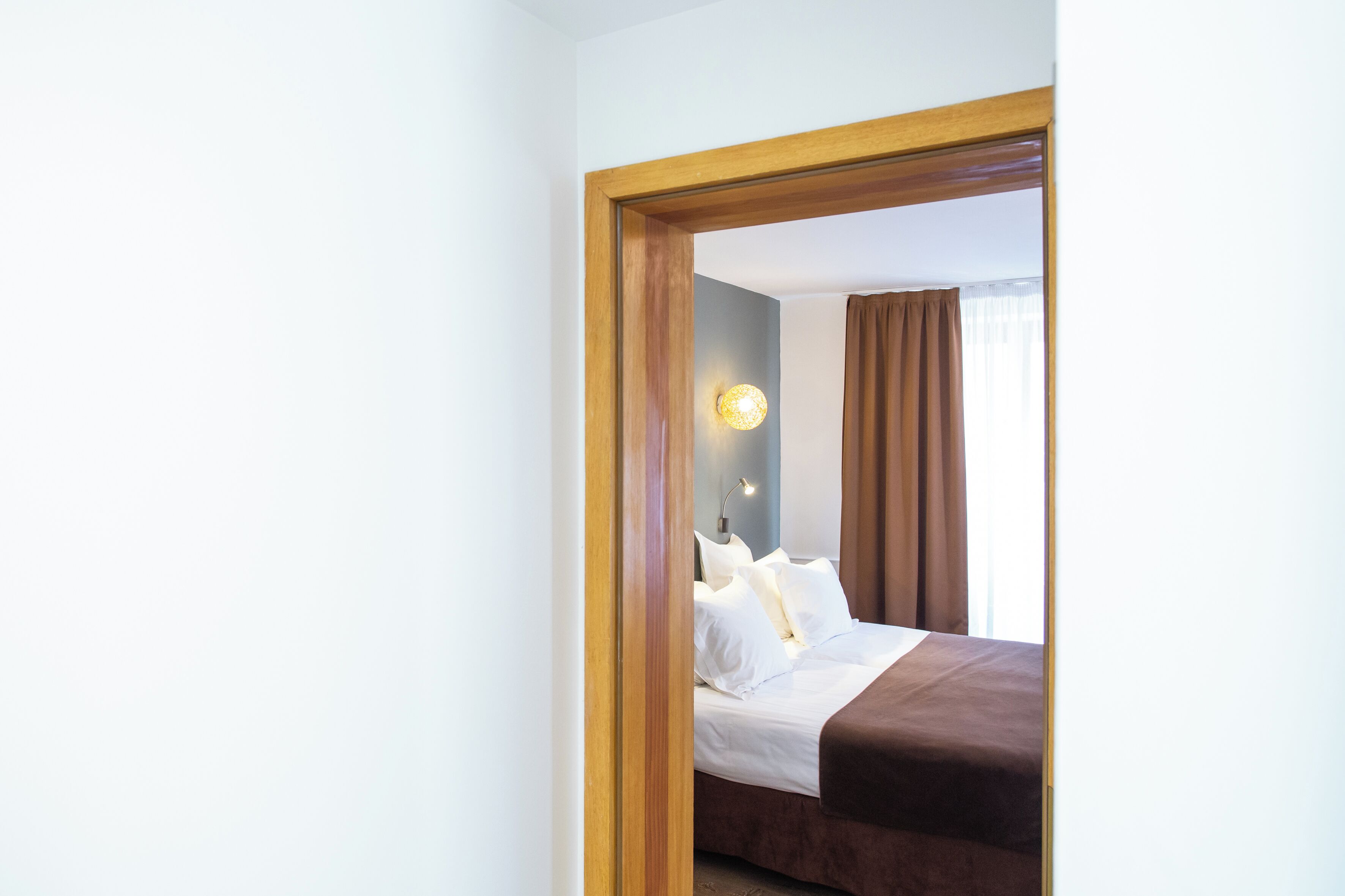 A hotel room with a bed, a nightstand, a wall-mounted lamp, and a window with curtains.