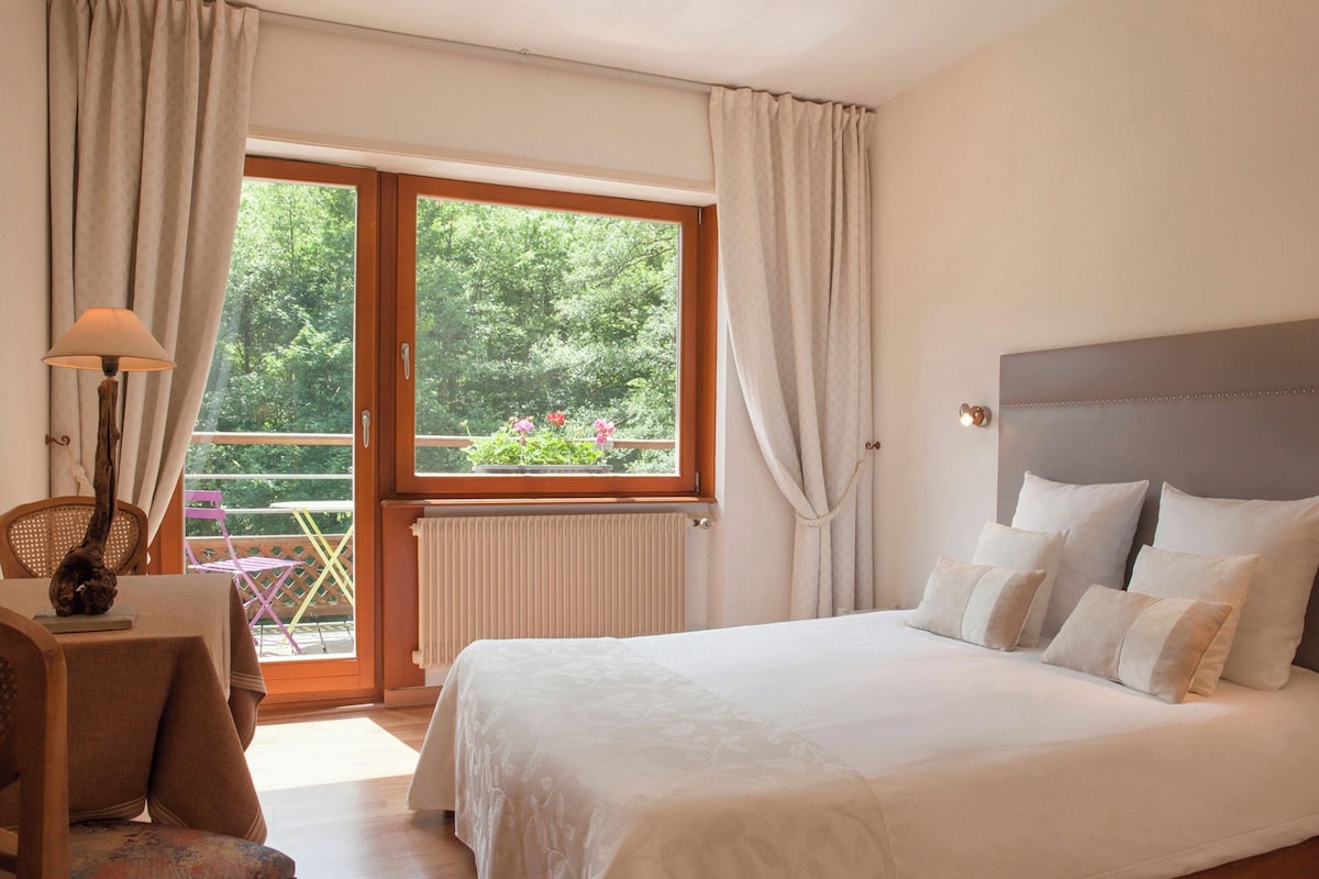 Une chambre à coucher avec un lit, une table de chevet, une lampe, une fenêtre avec des rideaux et une vue sur la verdure à l’extérieur.