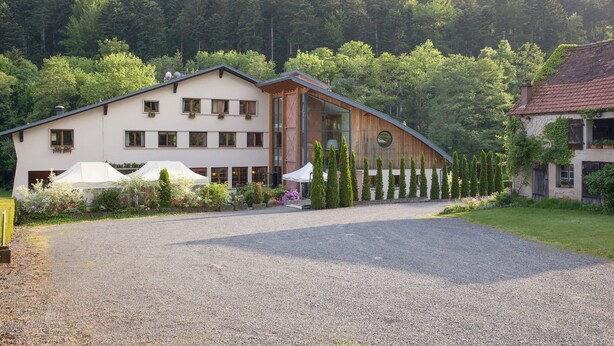 A two-story building with a gravel driveway, surrounded by greenery and a forested hillside. - La Fischhutte, The Originals Relais (Mollkirch)