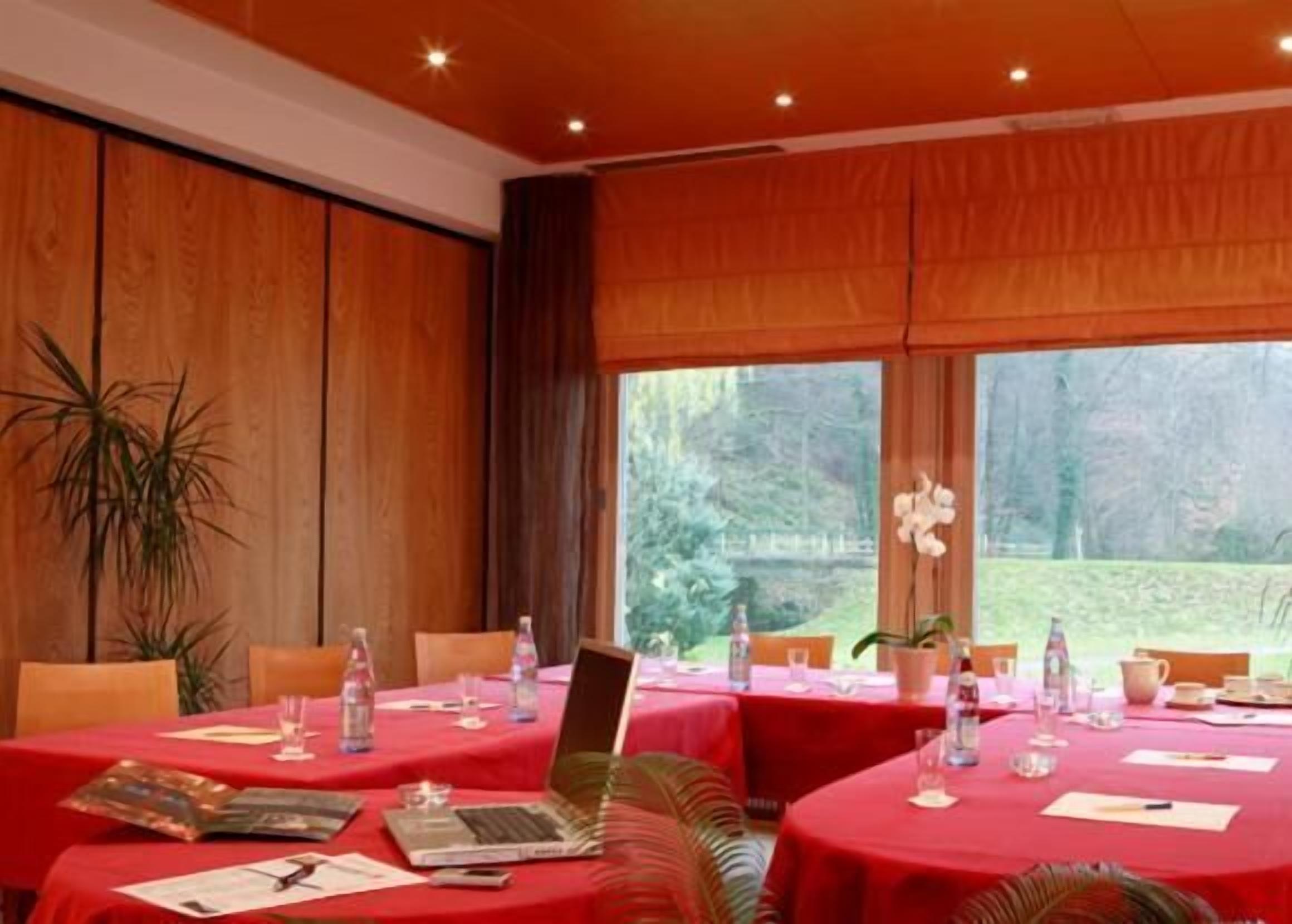 Un salón de conferencias con mesas redondas cubiertas con manteles rojos, equipado con computadoras portátiles, botellas de agua y documentos.