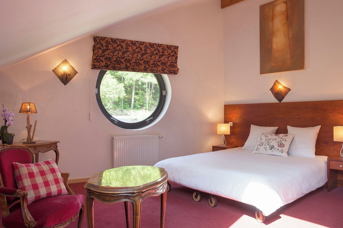 Une chambre avec un grand lit, une fenêtre ronde, un radiateur, une chaise, une petite table et deux lampes fixées au mur.