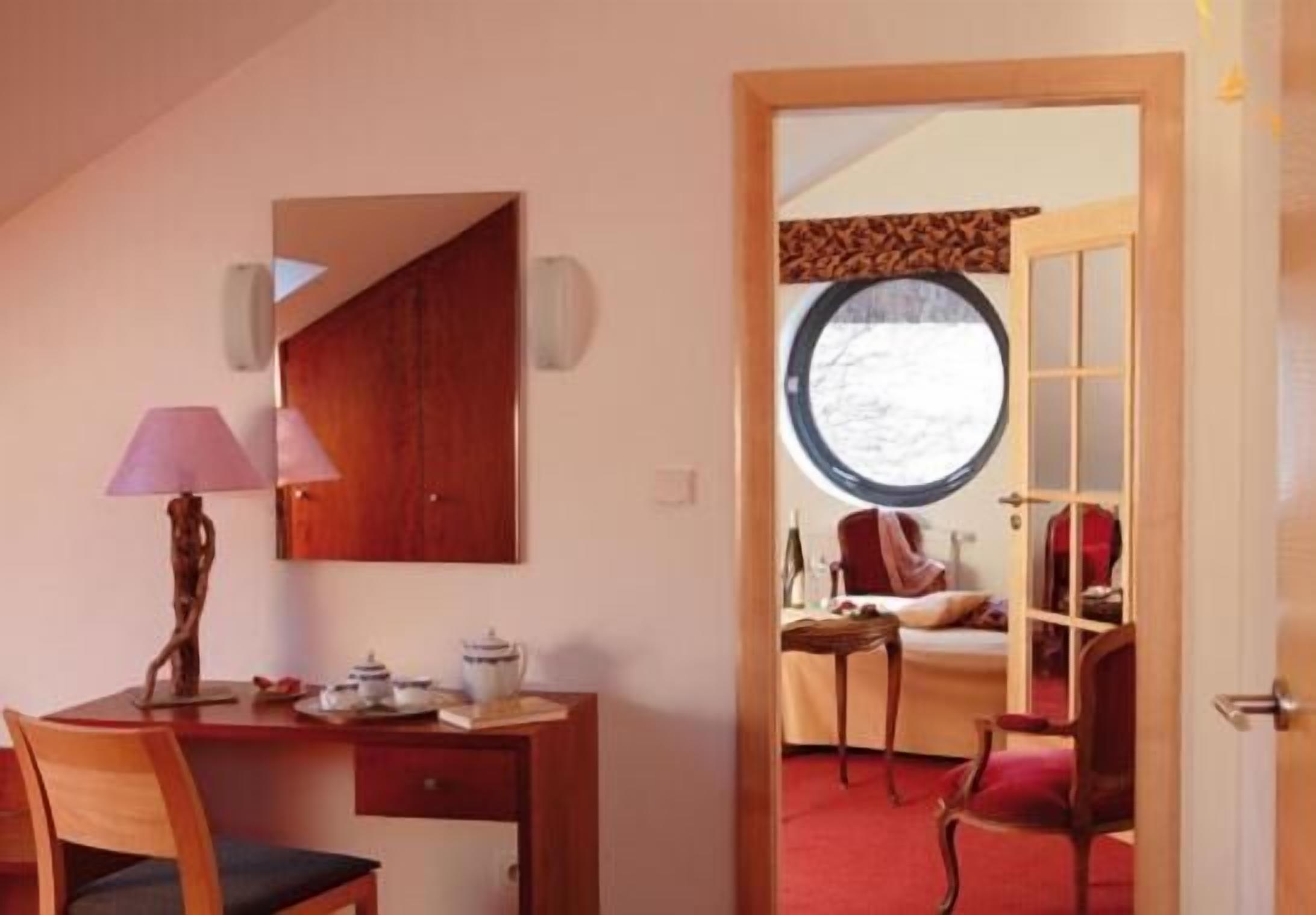 Habitación con escritorio, silla y cama sobre alfombra roja. Se ve una ventana con vistas a árboles y una puerta que da a otra habitación.