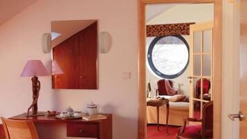 Habitación con escritorio, silla y cama sobre alfombra roja. Se ve una ventana con vistas a árboles y una puerta que da a otra habitación.