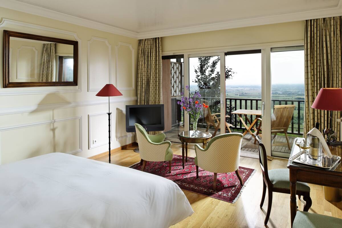 Habitación de hotel con cama, dos sillones, una mesita con un florero, televisor y balcón con vistas.