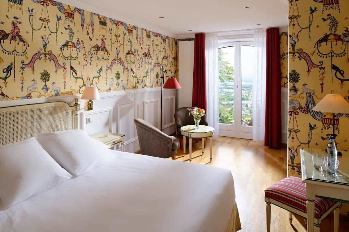 Habitación de hotel con una cama grande, una silla, una mesita con un florero y una ventana con cortinas.