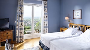 A bedroom with two beds, a television, a desk, a chair, a table with glasses and a bottle, and a view through the window. - Le Clos Saint Vincent (Ribeauvillé)