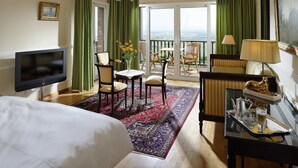 A hotel room with a large bed, a flat-screen TV, a dining area with chairs and a table, a balcony with a view, and a decorative lamp. - Le Clos Saint Vincent (Ribeauvillé)