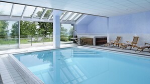 A modern indoor swimming pool with clear glass walls, wooden lounge chairs, and a stone fireplace. - Le Clos Saint Vincent (Ribeauvillé)
