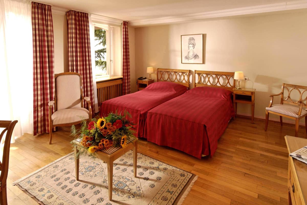 Habitación de hotel con dos camas, una silla de madera, una mesita con un arreglo floral y un cuadro en la pared.