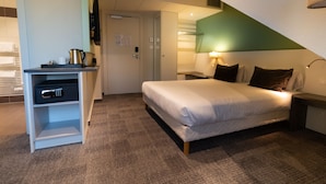 A hotel room with a bed, a desk with a safe, a lamp, and a view of the hallway. - greet Hôtel Colmar (Colmar)