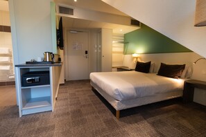 A hotel room with a bed, a desk with a safe, a lamp, and a view of the hallway. - greet Hôtel Colmar (Colmar)