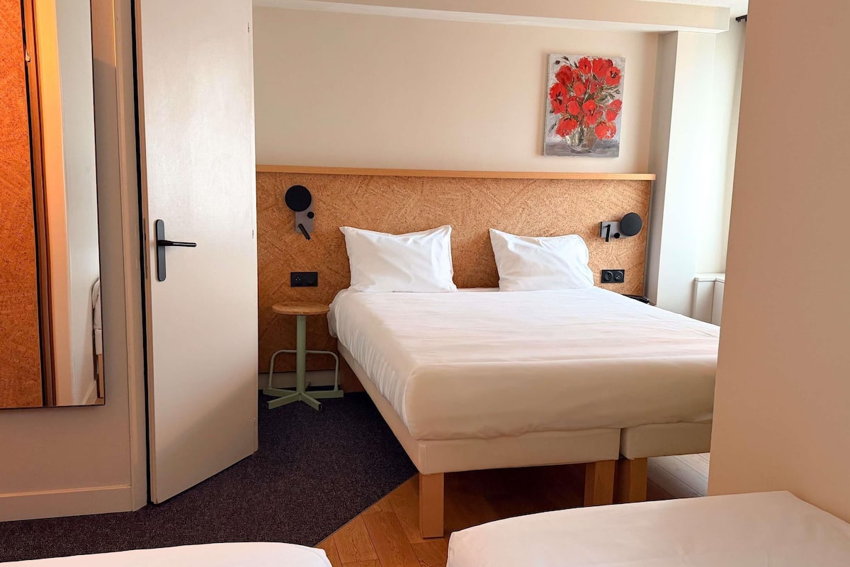 A hotel room with a bed, a small table, a chair, a wall clock, and a painting.