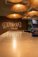 Zona de recepción con un cartel que dice 'greet', lámparas colgantes de mimbre, un mostrador de recepción y un florero con bolígrafos.