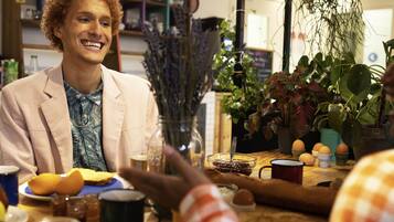 Une personne portant une veste de couleur claire est assise à une table devant divers plats, une tasse de café et un verre avec une cuillère.