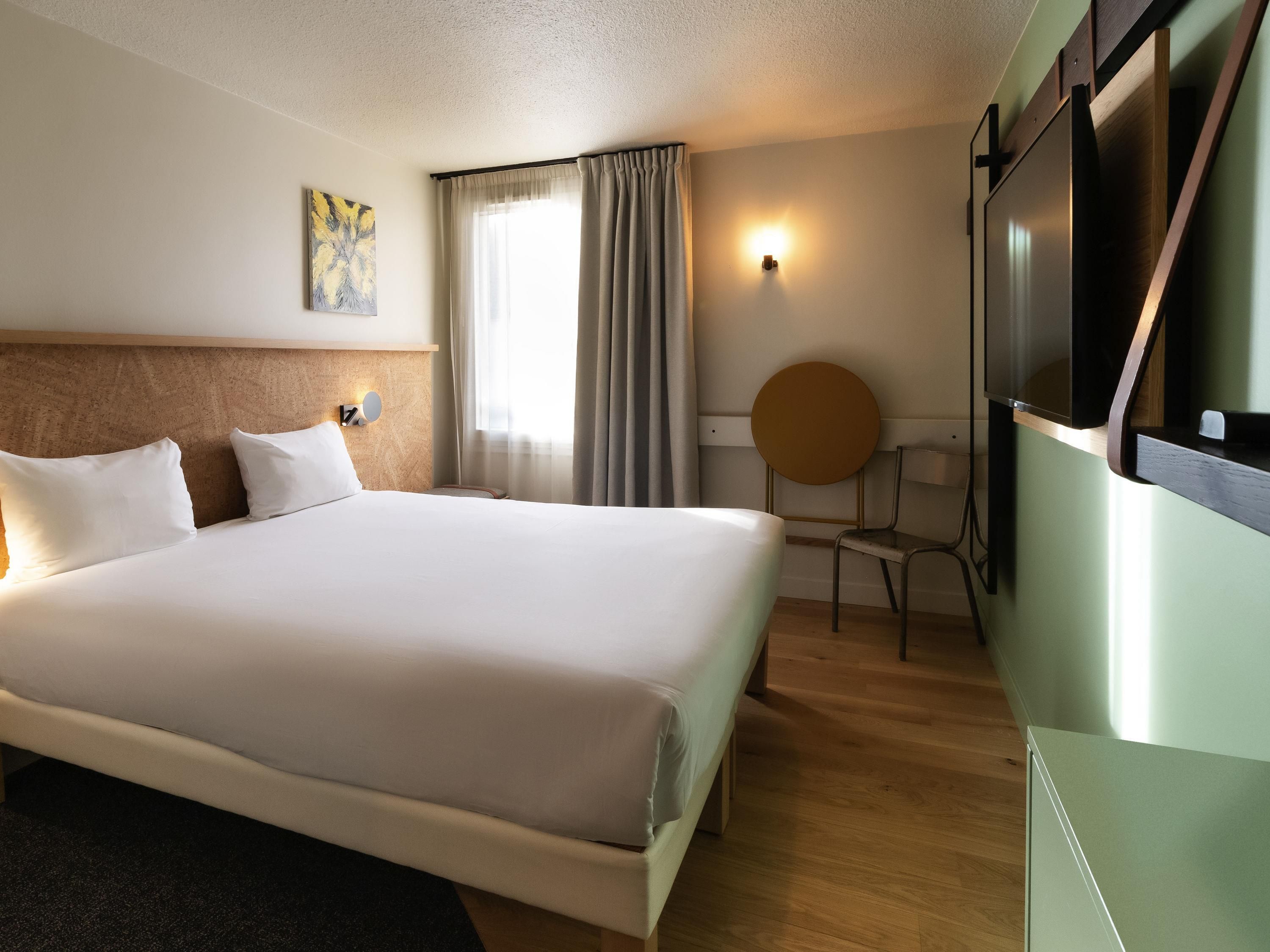 A hotel room with a large bed, a desk, a chair, a TV, and a window with curtains.