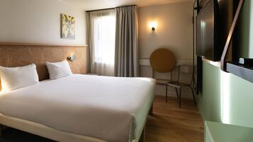A hotel room with a large bed, a desk, a chair, a TV, and a window with curtains.