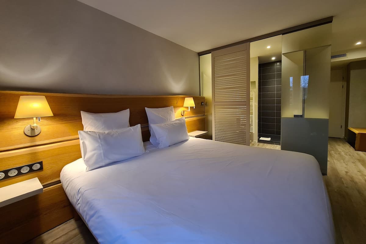 Et moderne hotelværelse med en stor seng, to sengeborde med lamper, et træhovedgærde og en skydedør til et badeværelse.