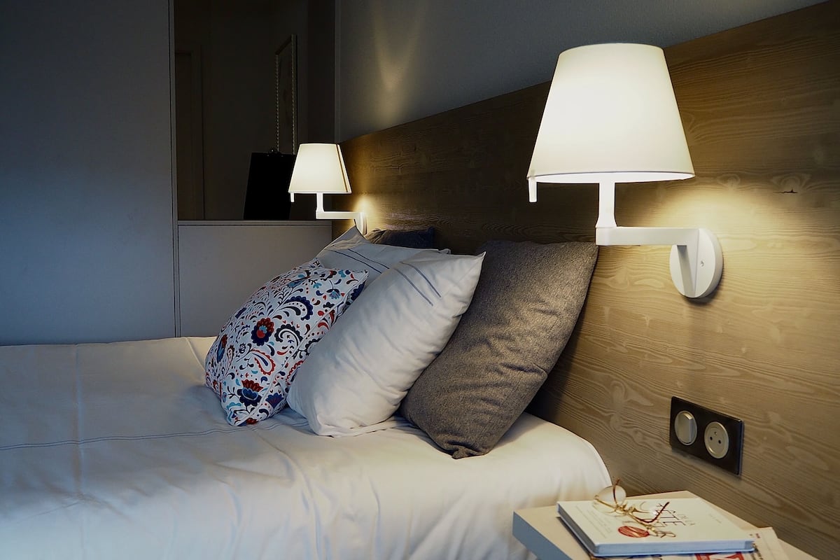 En pænt redt seng med mønstrede puder, et sengebord med bog og briller, samt vægmonterede lamper.