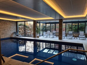 A modern indoor swimming pool with clear glass walls, lounge chairs, and a stone wall. - Hôtel Jenny (Hagenthal-le-Bas)