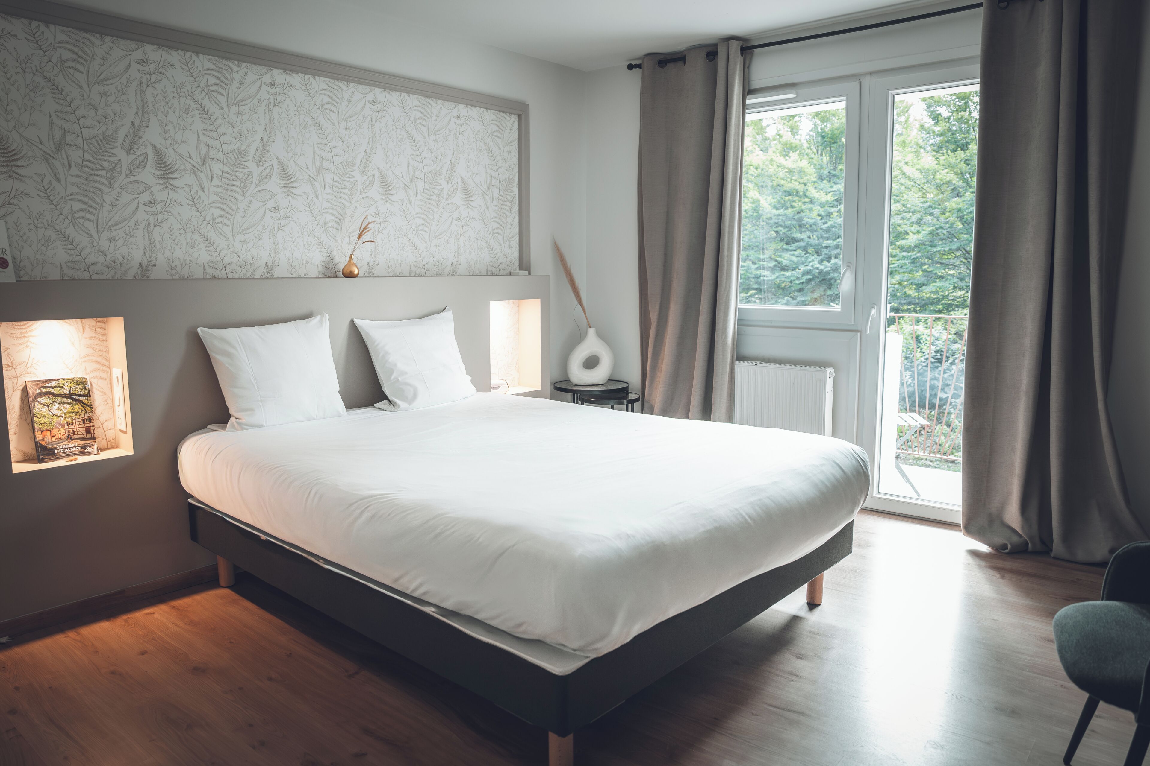 Une chambre avec un grand lit, une table de chevet, un vase et une fenêtre avec des rideaux.