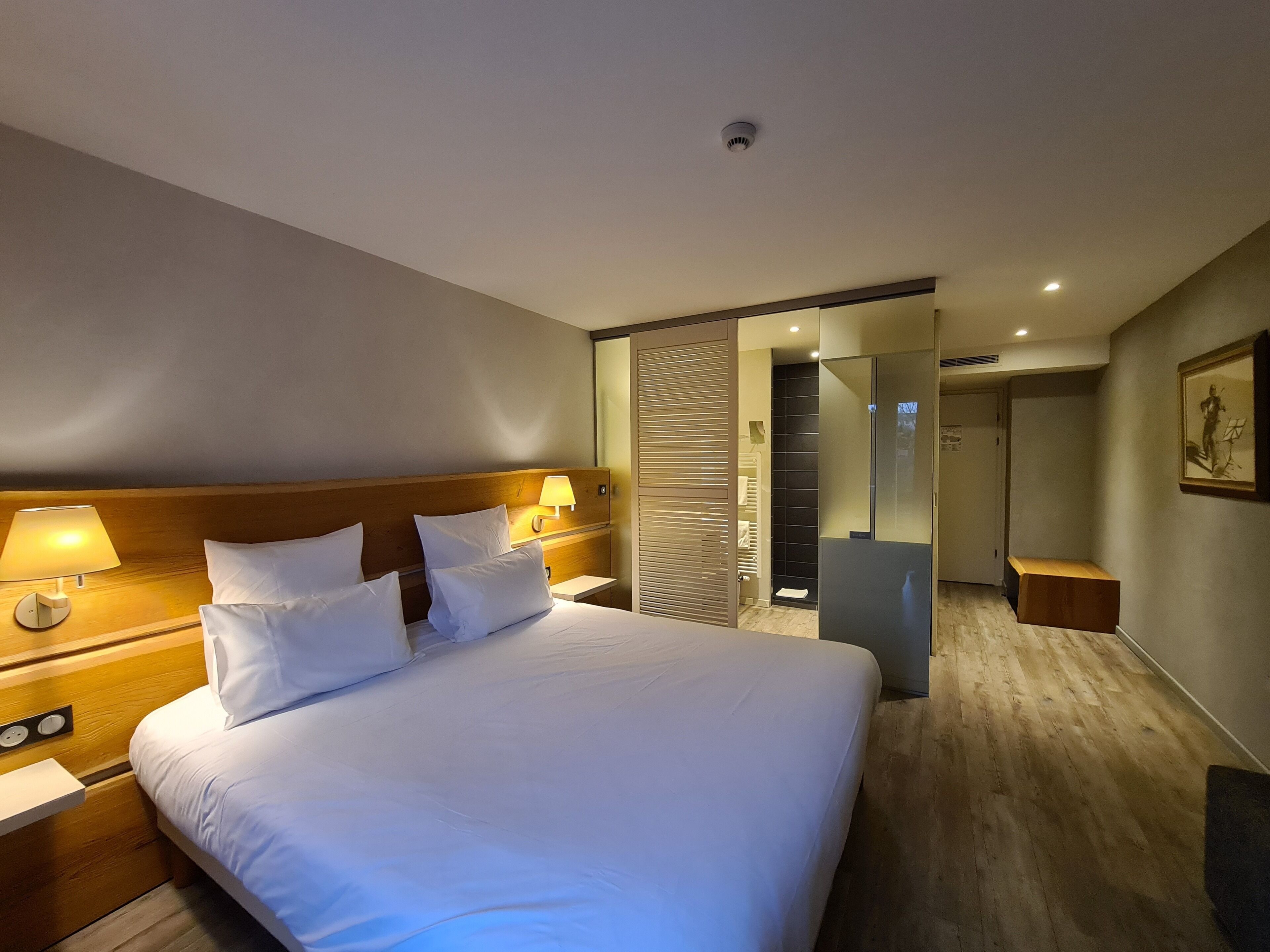 Una habitación de hotel con una cama grande, lámparas de noche, un cabecero de madera, un vestidor amplio y un cuadro enmarcado en la pared.