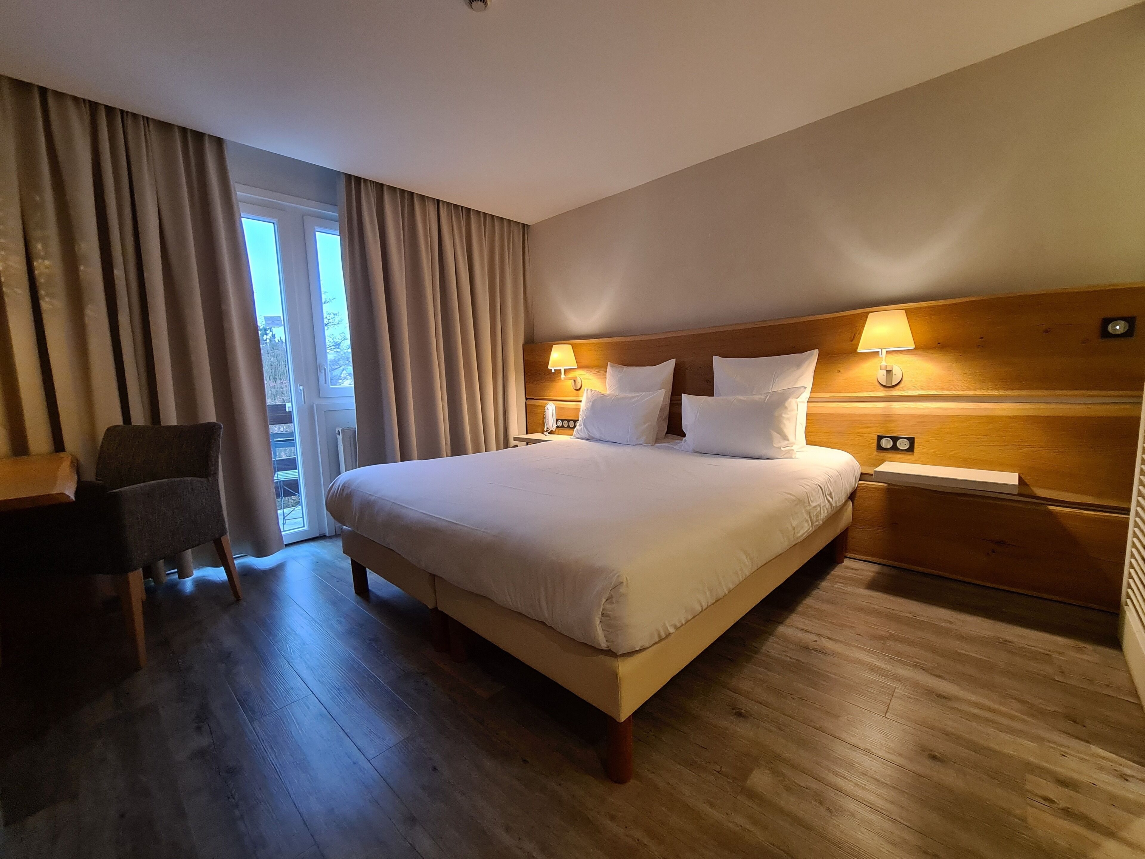 A hotel room with a large bed, two bedside lamps, a chair, and a view of the outdoors through a window with curtains.