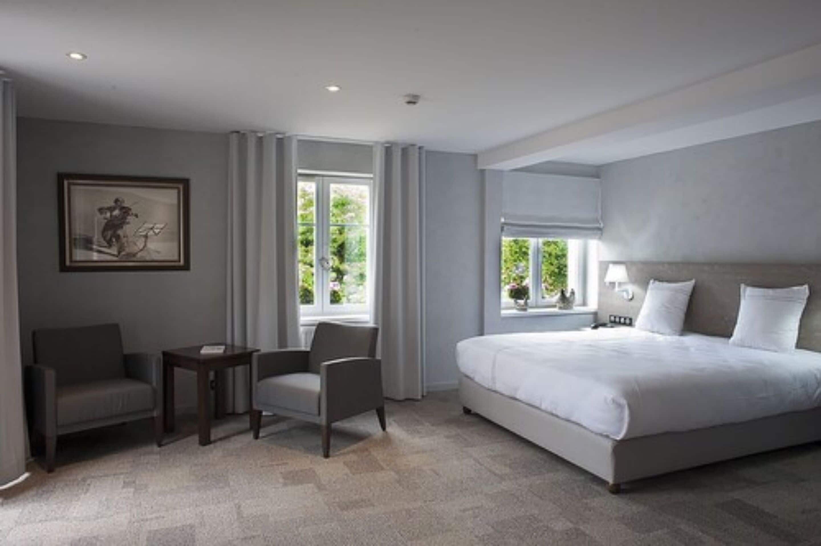 Ein modernes Hotelzimmer mit einem großen Bett, zwei Sesseln, einem kleinen Tisch und einem Fenster mit Vorhängen.