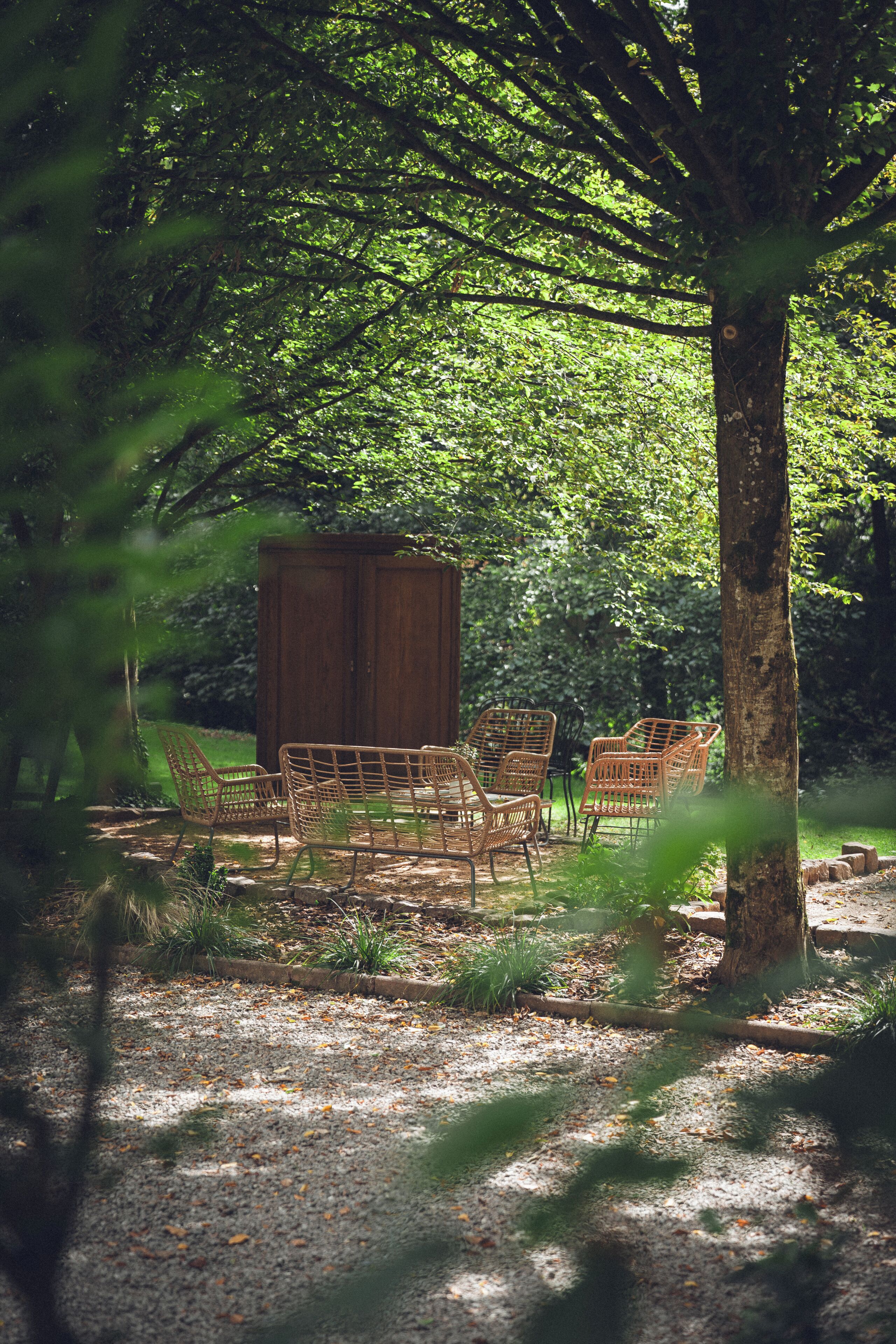 Een terras met rieten stoelen en een houten kast, omgeven door bomen en begroeiing.