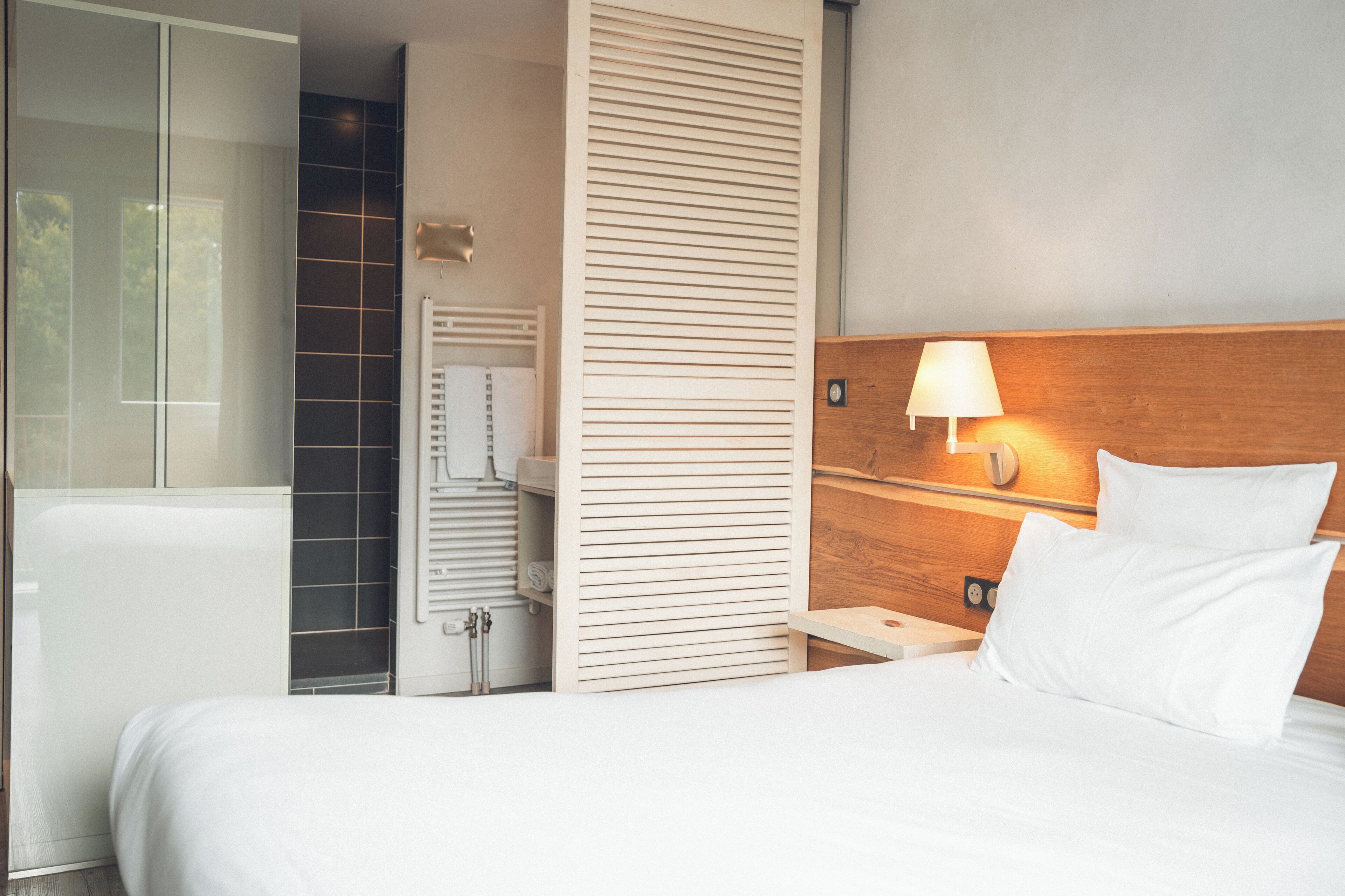 Een hotelkamer met een bed, nachtkastje, wandlamp, houten hoofdbord en een badkamer met douche en handdoekhouder.