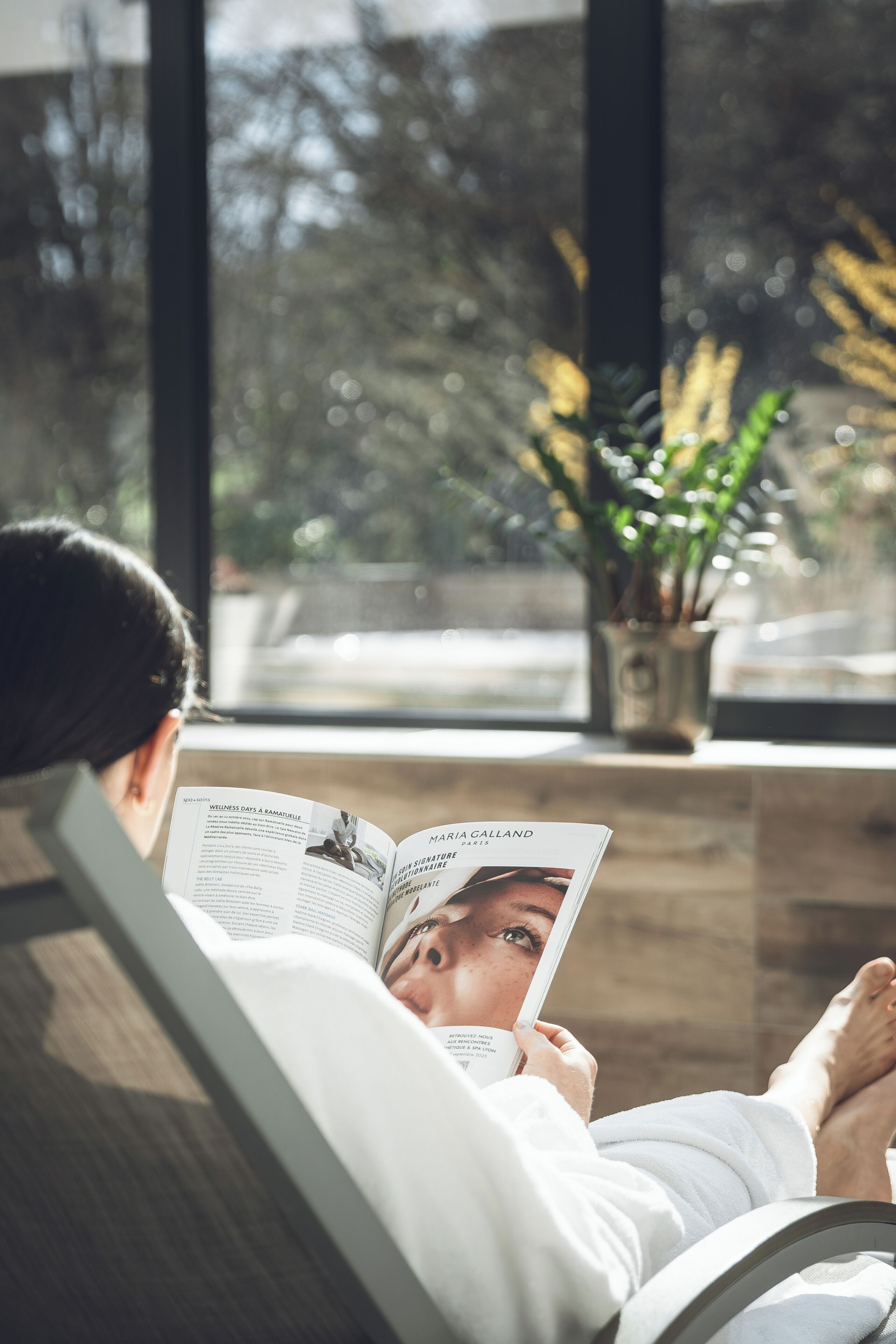 Una persona leyendo una revista en una habitación con un ventanal grande, una planta y vistas a árboles.