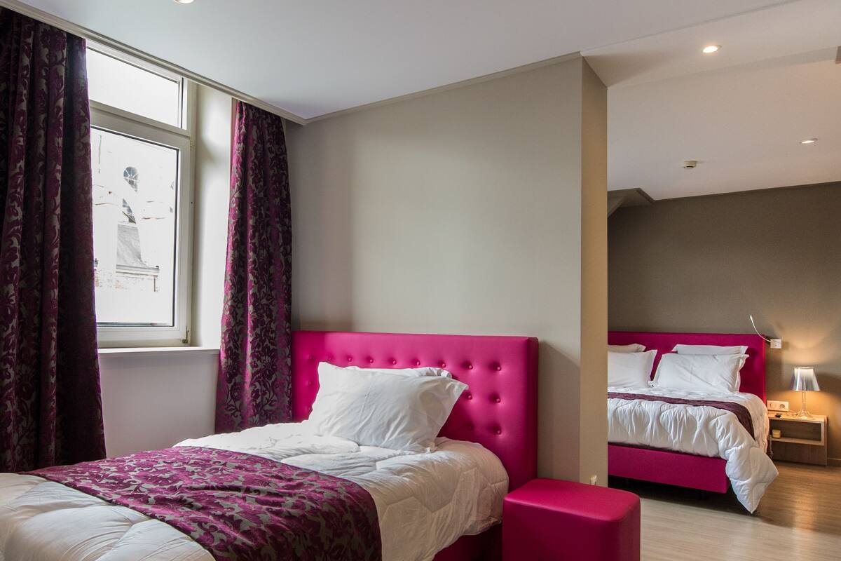 A hotel room with a large bed, a smaller bed, and a pink ottoman.