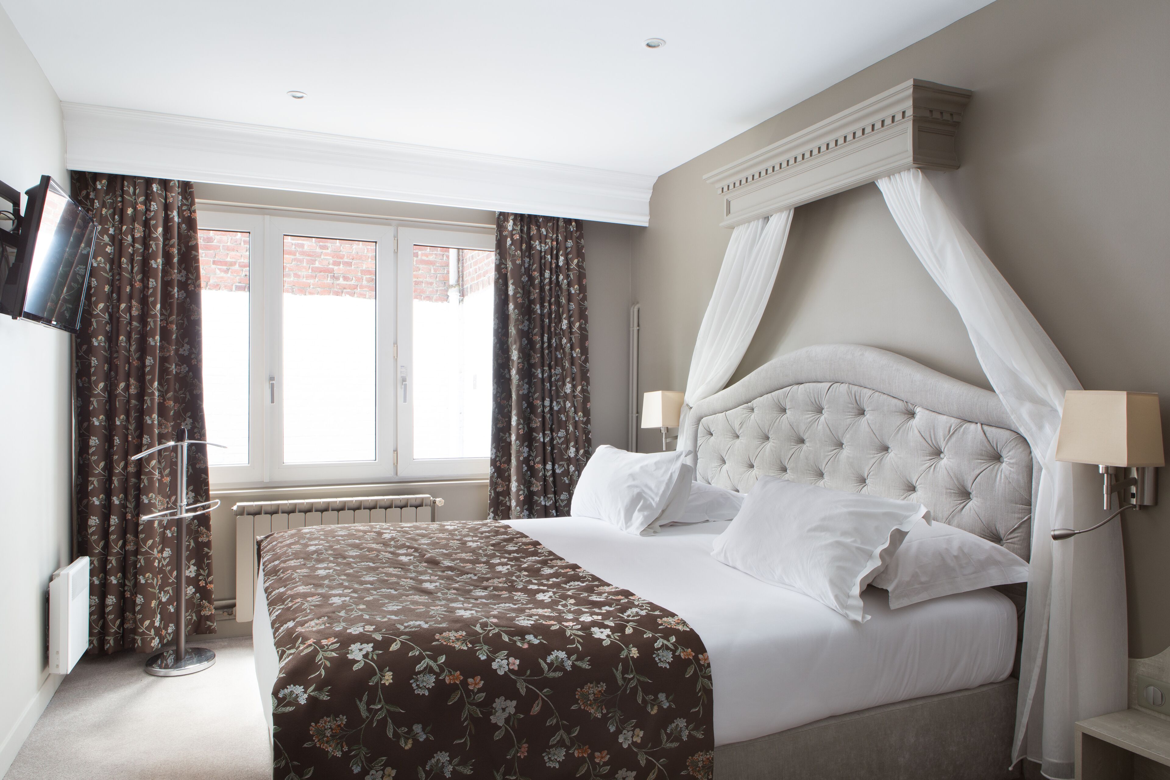 A neatly made bed with a floral bedspread, a tufted headboard, and a canopy. There is a window with curtains, a radiator, and a wall-mounted TV.
