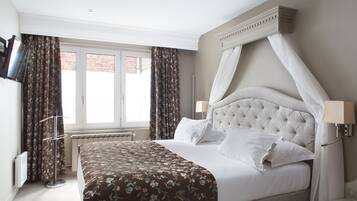A neatly made bed with a floral bedspread, a tufted headboard, and a canopy. There is a window with curtains, a radiator, and a wall-mounted TV.