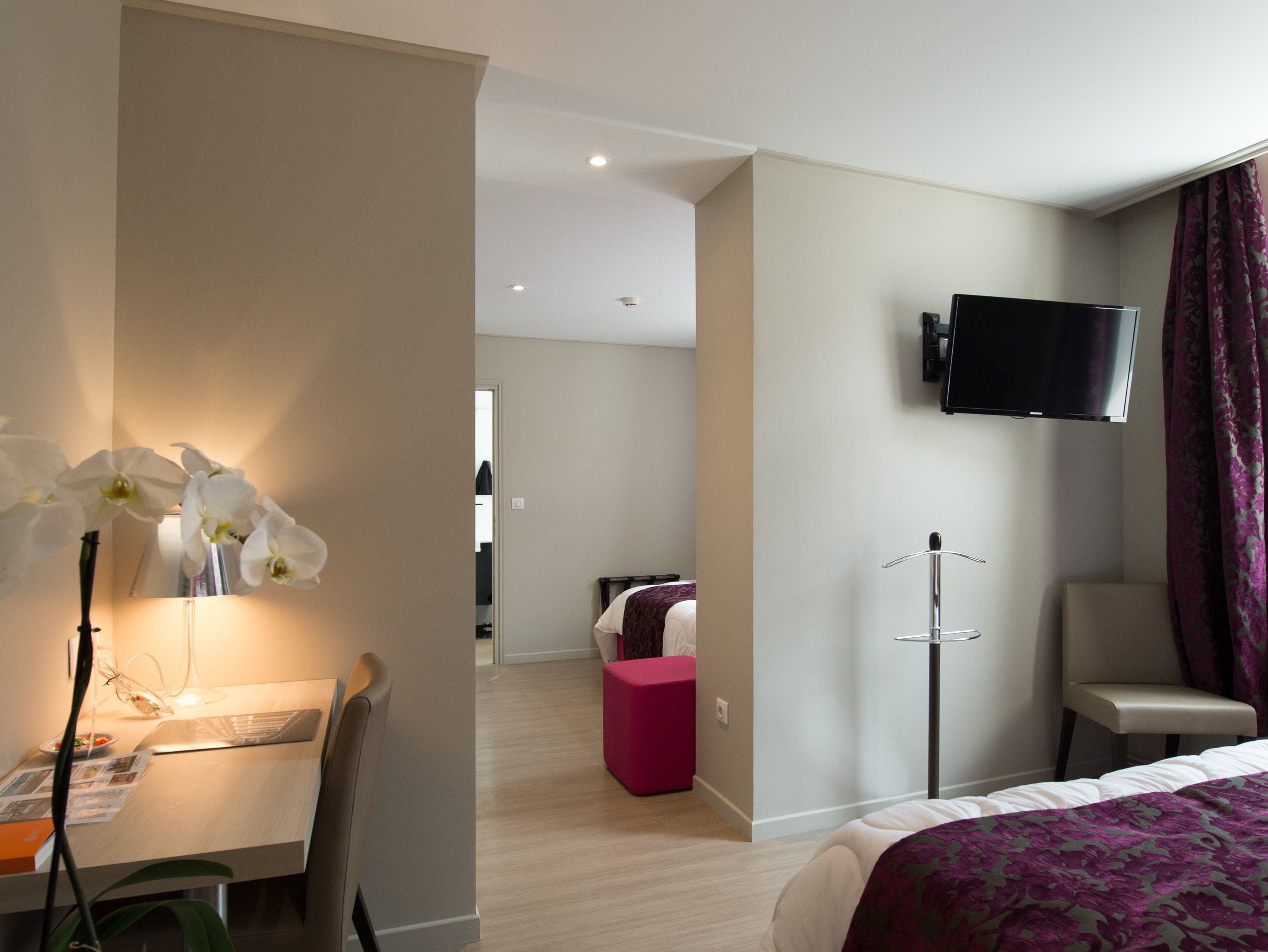 Ein Hotelzimmer mit einem Bett, einem Schreibtisch mit Stuhl, einem an der Wand befestigten Fernseher und einem kleinen rosa Hocker.