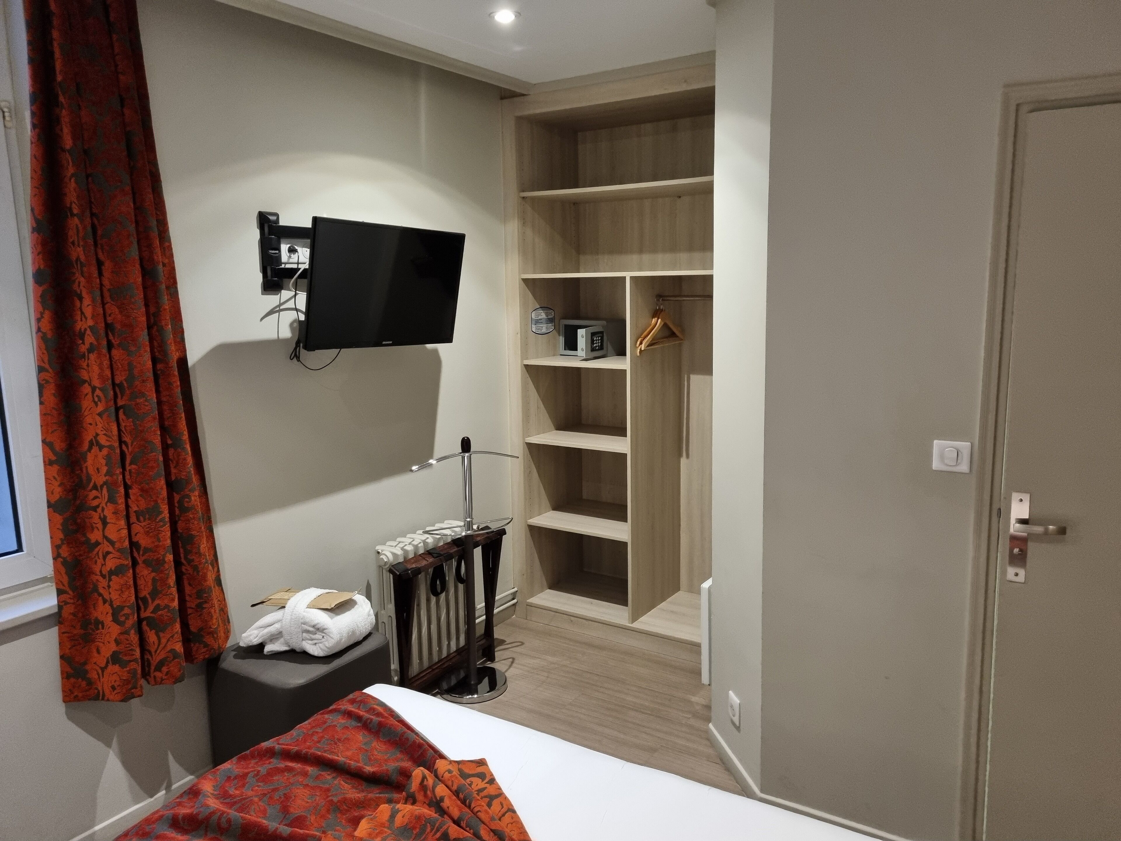 Habitación de hotel con cama, televisión montada en la pared, armario con estantes y una mesita con toalla y control remoto.